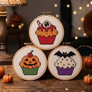 Puede incluir: Tres bastidores de bordado circulares de madera con dise&ntilde;os de punto de cruz de cupcakes. El bastidor superior muestra un cupcake rojo con un globo ocular. El inferior izquierdo muestra un cupcake de calabaza naranja con una cereza. El inferior derecho muestra un cupcake morado con un murci&eacute;lago.