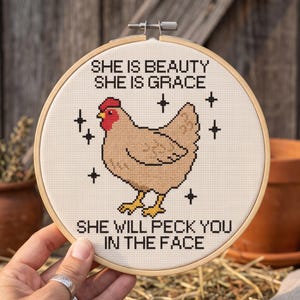 Puede incluir: Un bastidor de bordado circular de madera con un dise&ntilde;o de punto de cruz de una gallina de color marr&oacute;n claro con cresta roja. El texto dice: SHE IS BEAUTY SHE IS GRACE SHE WILL PECK YOU IN THE FACE. Acentos negros en forma de estrella rodean a la gallina.