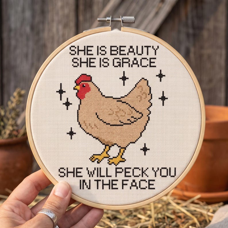 Puede incluir: Un bastidor de bordado circular de madera con un dise&ntilde;o de punto de cruz de una gallina de color marr&oacute;n claro con cresta roja. El texto dice: SHE IS BEAUTY SHE IS GRACE SHE WILL PECK YOU IN THE FACE. Acentos negros en forma de estrella rodean a la gallina.