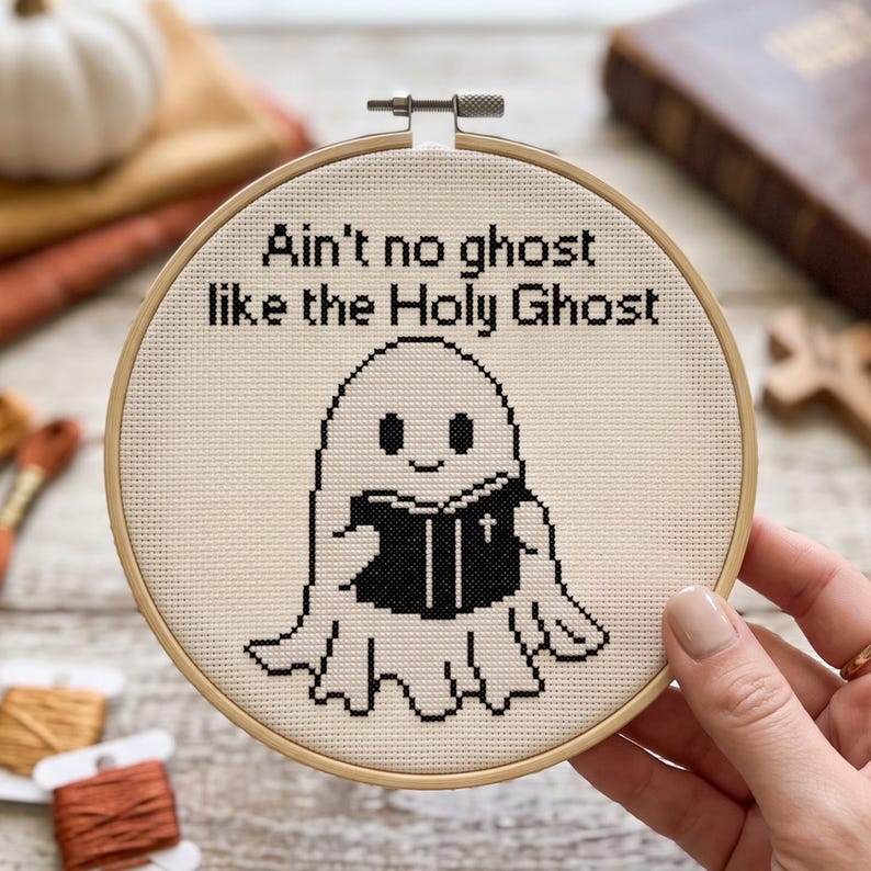 May include: A circular wooden embroidery hoop containing a cross-stitch design on cream fabric. The design features a black ghost holding a black book with a white cross on the cover, with the text "Ain't no ghost like the Holy Ghost" above.