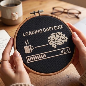 May include: A circular wooden embroidery hoop containing black fabric with a cross-stitch design. The design features the text LOADING CAFFEINE, a coffee mug icon, a brain icon, and a progress bar showing 42% completion in light beige thread.