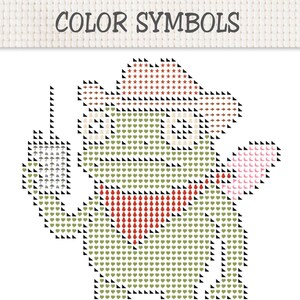 May include: A cross-stitch pattern titled COLOR SYMBOLS featuring a green frog wearing a cowboy hat and a red bandana. The frog holds a small rectangular sign with umbrella icons. The design uses various symbols like hearts and stars.