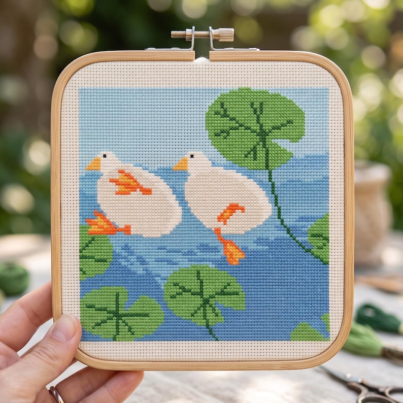 K&ouml;nnte beinhalten: Ein quadratischer Stickrahmen aus Holz mit einem Kreuzstichmuster von zwei wei&szlig;en Enten mit orangefarbenen Schn&auml;beln und F&uuml;&szlig;en, die auf blauem Wasser schwimmen. Die Szene zeigt mehrere gr&uuml;ne Seerosenbl&auml;tter auf dem Wasser vor einem hellblauen Himmel.