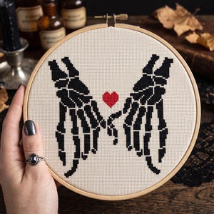 May include: A circular wooden embroidery hoop containing a cross-stitch pattern on cream-colored fabric. The design features two black skeleton hands reaching toward each other with fingers touching, centered around a small, solid red heart.