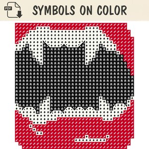 Op de afbeelding: Een kruissteekpatroon getiteld SYMBOLS ON COLOR met een centrale zwarte vleermuissilhouet tegen een witte cirkelvormige achtergrond, geplaatst op een rood raster. Het ontwerp gebruikt herhalende pijlsymbolen voor de steekplaatsing.
