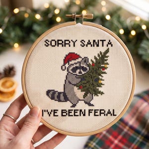 May include: A circular wooden embroidery hoop containing a cross-stitch design on cream fabric. The design features a grey raccoon wearing a red Santa hat and holding a green Christmas tree, with the black text SORRY SANTA I'VE BEEN FERAL.