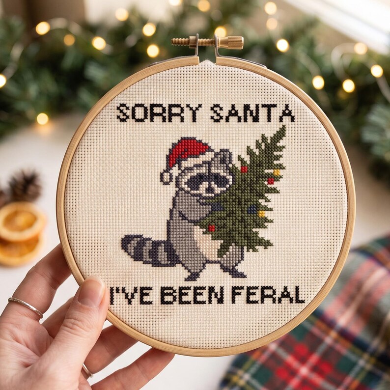 May include: A circular wooden embroidery hoop containing a cross-stitch design on cream fabric. The design features a grey raccoon wearing a red Santa hat and holding a green Christmas tree, with the black text SORRY SANTA I'VE BEEN FERAL.