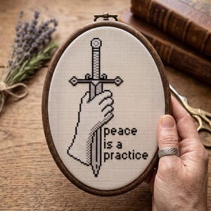 K&ouml;nnte beinhalten: Ein ovaler Stickrahmen mit einem Kreuzstichmotiv auf cremefarbenem Stoff. Das Design zeigt eine Hand, die ein Schwert in Grau- und Schwarzt&ouml;nen h&auml;lt, mit dem Text "peace is a practice", der in schwarzen Buchstaben rechts neben dem Schwert gestickt ist.