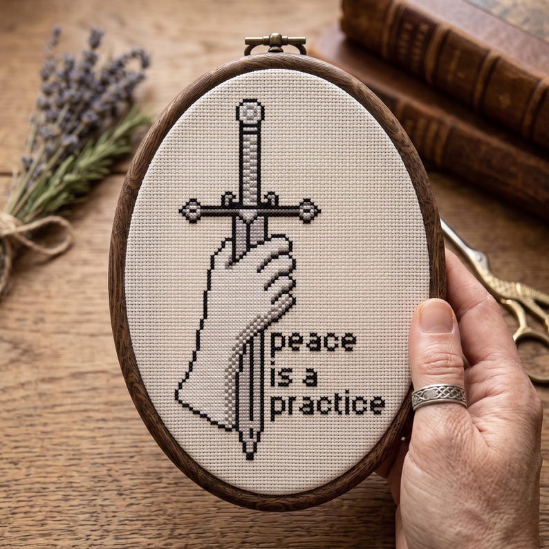 K&ouml;nnte beinhalten: Ein ovaler Stickrahmen mit einem Kreuzstichmotiv auf cremefarbenem Stoff. Das Design zeigt eine Hand, die ein Schwert in Grau- und Schwarzt&ouml;nen h&auml;lt, mit dem Text "peace is a practice", der in schwarzen Buchstaben rechts neben dem Schwert gestickt ist.