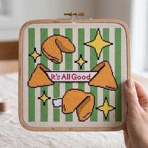 May include: A square wooden embroidery hoop containing a cross-stitch pattern on white fabric with vertical green stripes. The design features three orange fortune cookies and yellow four-pointed stars, with a banner reading It's All Good.