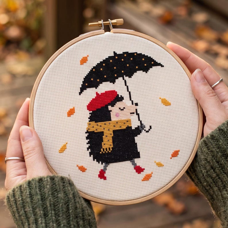 May include: A circular wooden embroidery hoop containing a cross-stitch design of a hedgehog wearing a red beret, a yellow scarf, and red boots. The hedgehog holds a black umbrella with yellow dots, surrounded by falling orange and yellow leaves.