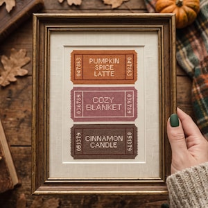 May include: A framed cross-stitch embroidery featuring three rectangular ticket designs in orange, mauve, and brown. The tickets display the text PUMPKIN SPICE LATTE, COZY BLANKET, and CINNAMON CANDLE, each with unique serial numbers on the sides.