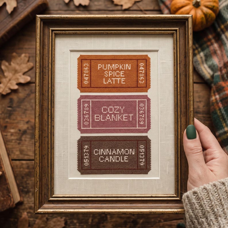 May include: A framed cross-stitch embroidery featuring three rectangular ticket designs in orange, mauve, and brown. The tickets display the text PUMPKIN SPICE LATTE, COZY BLANKET, and CINNAMON CANDLE, each with unique serial numbers on the sides.