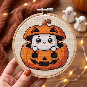 May include: A cross-stitch embroidery piece in a round wooden hoop featuring a white ghost character peeking out from inside a carved orange jack-o'-lantern pumpkin. The design uses orange, black, and white thread on a cream-colored fabric background.