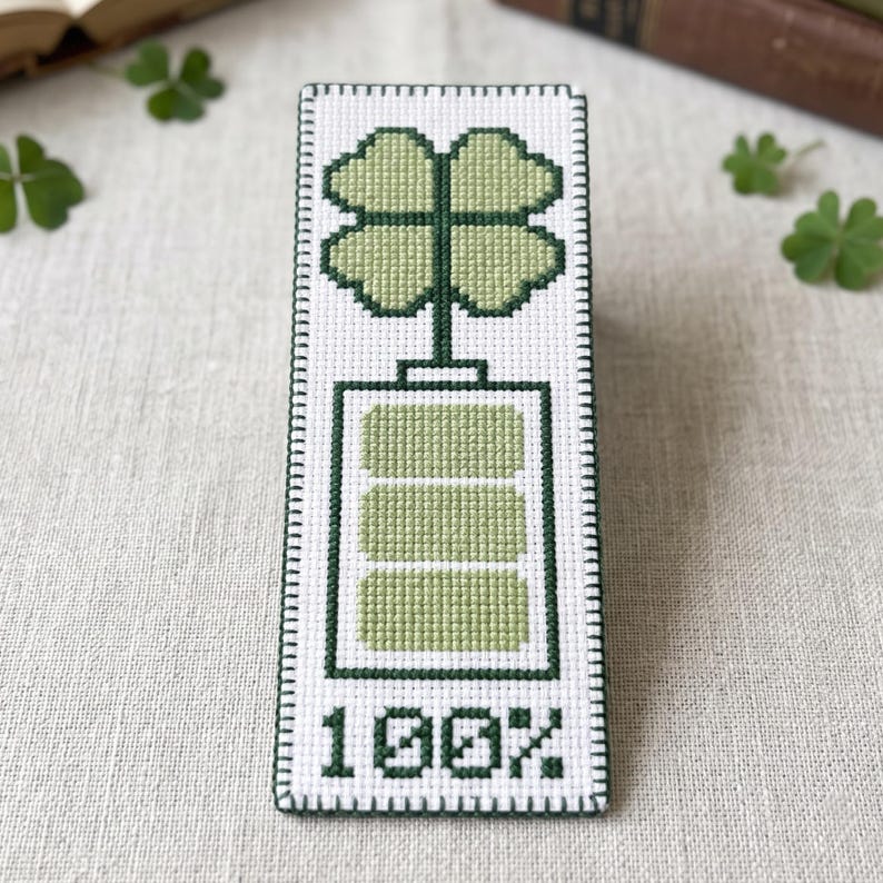 May include: A rectangular cross-stitch piece on white fabric featuring a dark green border. The design displays a battery icon filled with light green segments, topped with a four-leaf clover, and the text 100% in dark green at the bottom.