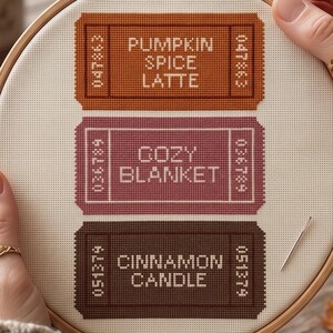May include: A cross-stitch embroidery project in a wooden hoop featuring three rectangular ticket-shaped designs. The top ticket is orange with the text PUMPKIN SPICE LATTE, the middle is mauve with COZY BLANKET, and the bottom is brown with CINNAMON CANDLE.