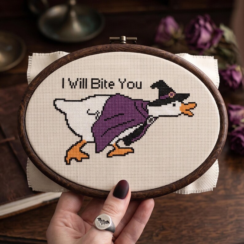 Op de afbeelding: Een ovale houten borduurring met een kruissteekontwerp op cr&egrave;mekleurige stof. Het ontwerp toont een witte gans met een paarse cape en een zwarte heksenhoed met een klein bloemetje. De tekst "I Will Bite You" is in het zwart geborduurd.