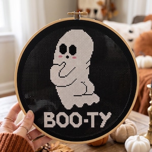 Puede incluir: Un bastidor de bordado circular de madera que contiene un fondo de tela negra con un fantasma en punto de cruz blanco. El fantasma tiene ojos negros grandes y mejillas rosadas. Debajo, el texto BOO-TY est&aacute; bordado en letras blancas.