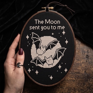Puede incluir: Un bastidor de bordado ovalado de madera que contiene tela negra con un dise&ntilde;o de punto de cruz. El dise&ntilde;o presenta una gran luna p&aacute;lida, dos peque&ntilde;os murci&eacute;lagos y varias estrellas en hilo blanco. El texto "The Moon sent you to me" est&aacute; bordado.