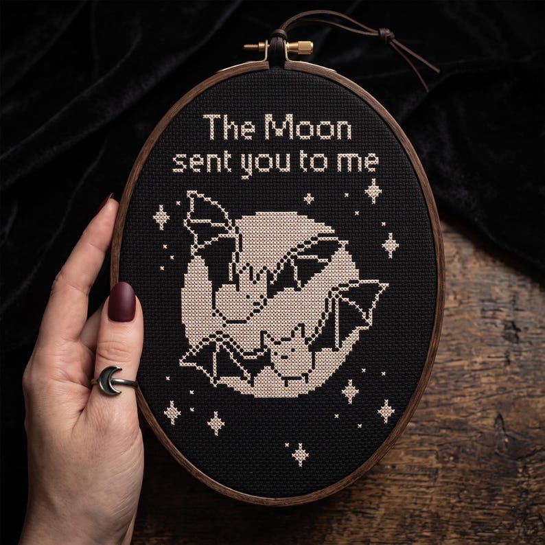 Puede incluir: Un bastidor de bordado ovalado de madera que contiene tela negra con un dise&ntilde;o de punto de cruz. El dise&ntilde;o presenta una gran luna p&aacute;lida, dos peque&ntilde;os murci&eacute;lagos y varias estrellas en hilo blanco. El texto "The Moon sent you to me" est&aacute; bordado.