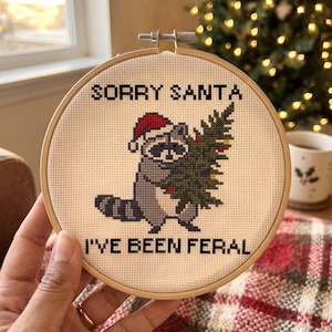 May include: A circular wooden embroidery hoop containing a cross-stitch design on white fabric. The design features a grey raccoon wearing a red Santa hat and holding a green Christmas tree with small ornaments. Text reads: SORRY SANTA I'VE BEEN FERAL.