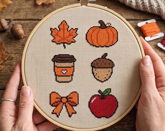 May include: A circular wooden embroidery hoop containing a beige fabric panel with six cross-stitched autumn motifs: an orange maple leaf, a pumpkin, a coffee cup with a heart, an acorn, an orange bow, and a red apple with a green leaf.