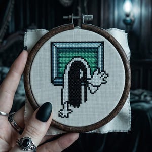 K&ouml;nnte beinhalten: Eine Kreuzstichstickerei in einem runden dunklen Holzrahmen mit einem verpixelten Motiv einer langhaarigen Figur, die aus einem gr&uuml;n get&ouml;nten Fernsehbildschirm hervorkommt. Die Figur hat schwarzes Haar, ein wei&szlig;es Gewand und erhobene H&auml;nde.