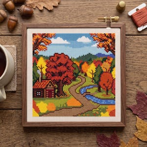 May include: A square wooden embroidery hoop containing a cross-stitch pattern of an autumn landscape. The scene features a small cabin, a winding dirt path, a blue stream, and colorful trees in shades of red, orange, and yellow under a blue sky.