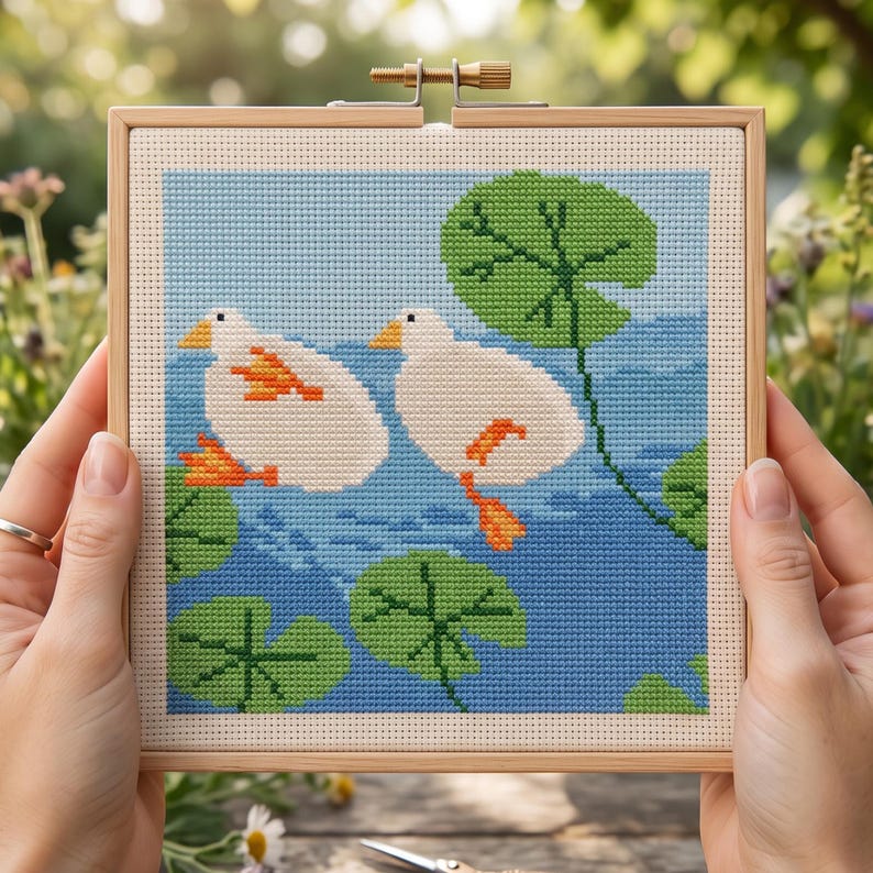 K&ouml;nnte beinhalten: Ein quadratisches Kreuzstich-Stickbild in einem hellen Holzrahmen. Das Motiv zeigt zwei wei&szlig;e Enten mit orangefarbenen Schn&auml;beln und F&uuml;&szlig;en, die auf einem blauen Wasserhintergrund schwimmen, umgeben von mehreren gr&uuml;nen Seerosenbl&auml;ttern.
