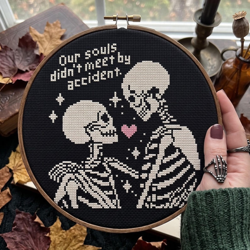Op de afbeelding: Een ronde houten borduurring met zwarte stof en een wit kruissteekontwerp van twee skeletten die tegenover elkaar staan. De tekst luidt, Our souls didn't meet by accident. Een klein roze hartje is tussen de twee skeletten geborduurd.