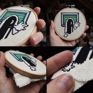 K&ouml;nnte beinhalten: Ein runder Stickrahmen aus Holz mit einer Kreuzsticharbeit auf wei&szlig;em Stoff. Das Motiv zeigt eine verpixelte schwarz-wei&szlig;e Figur mit ausgestreckten H&auml;nden, die aus einem gr&uuml;n-grauen, rechteckigen Fensterrahmen hervorkommt.