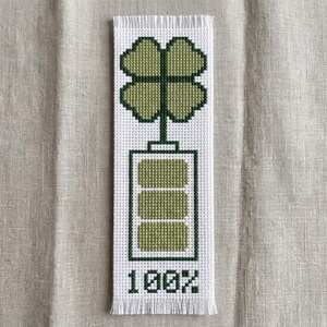May include: A rectangular white bookmark featuring a cross-stitch design of a four-leaf clover atop a battery icon with three filled green segments and the text 100% at the bottom, finished with a fringed edge at the top and bottom of the piece.