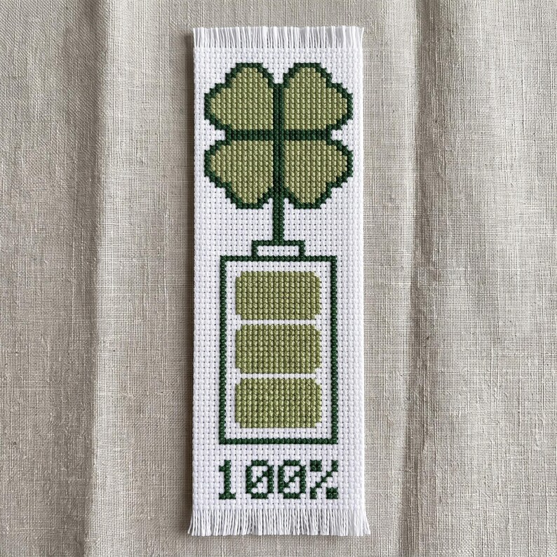 May include: A rectangular white bookmark featuring a cross-stitch design of a four-leaf clover atop a battery icon with three filled green segments and the text 100% at the bottom, finished with a fringed edge at the top and bottom of the piece.