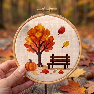May include: A circular wooden embroidery hoop containing a cross-stitch pattern on cream fabric. The design features a vibrant orange and yellow autumn tree, a brown park bench, a small orange pumpkin, and scattered fallen leaves in shades of red.