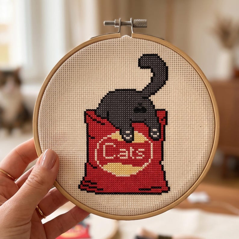 K&ouml;nnte beinhalten: Ein runder Stickrahmen aus Holz mit einem Kreuzstichmotiv, das eine schwarze Katze zeigt, die aus einer roten T&uuml;te schaut. Die T&uuml;te hat ein gelbes, kreisf&ouml;rmiges Emblem mit dem in wei&szlig;en Buchstaben gestickten Wort "Cats" auf beigem Stoff.