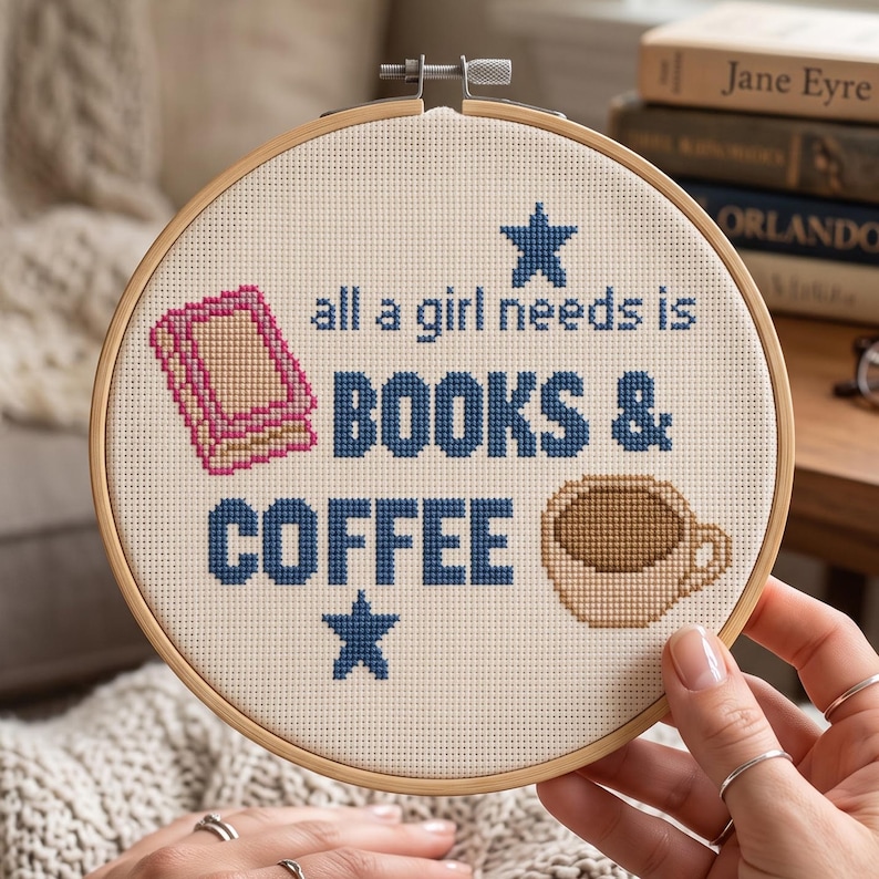 May include: A circular wooden embroidery hoop containing a cream-colored fabric with cross-stitched text that reads all a girl needs is BOOKS & COFFEE. The design includes a pink book icon, a brown coffee mug icon, and two dark blue stars.