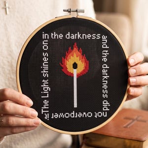 Puede incluir: Un bastidor de bordado circular de madera con tela negra y un dise&ntilde;o de punto de cruz de una cerilla encendida en rojo, naranja y amarillo. El texto blanco dice: The Light shines on in the darkness and the darkness did not overpower it.