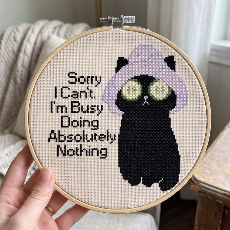 May include: A circular wooden embroidery hoop containing a cross-stitch design on cream fabric. The design features a black cat wearing a light purple towel wrap on its head and cucumber slices over its eyes, with the text Sorry I Can't. I'm BUSY Doing Absolutely Nothing.