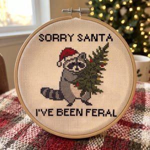 May include: A circular wooden embroidery hoop containing a cross-stitch design on white fabric. The design features a grey raccoon wearing a red Santa hat and holding a green Christmas tree with small ornaments. The text reads SORRY SANTA I'VE BEEN FERAL.