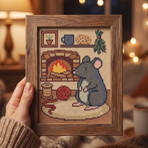 Pode incluir: Um bordado em ponto cruz emoldurado apresentando um rato cinzento sentado num tapete em frente a uma lareira. A cena inclui um carretel de linha, um novelo de l&atilde; vermelha, uma prateleira com uma caneca azul, bolachas, um pequeno quadro e ervas.