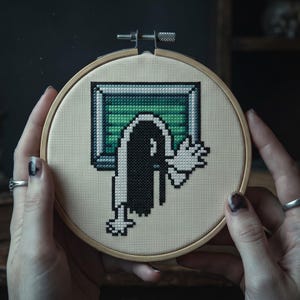 K&ouml;nnte beinhalten: Ein runder Stickrahmen aus Holz mit einem Kreuzstich-Motiv auf beigem Stoff. Das Bild im Pixel-Art-Stil zeigt eine Figur mit langem schwarzem Haar, die aus einem rechteckigen, gr&uuml;n-grauen Fernsehbildschirm mit ausgestreckten wei&szlig;en H&auml;nden hervorkommt.