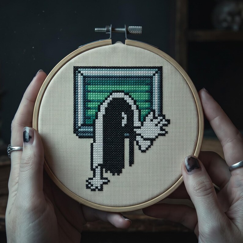 K&ouml;nnte beinhalten: Ein runder Stickrahmen aus Holz mit einem Kreuzstich-Motiv auf beigem Stoff. Das Bild im Pixel-Art-Stil zeigt eine Figur mit langem schwarzem Haar, die aus einem rechteckigen, gr&uuml;n-grauen Fernsehbildschirm mit ausgestreckten wei&szlig;en H&auml;nden hervorkommt.