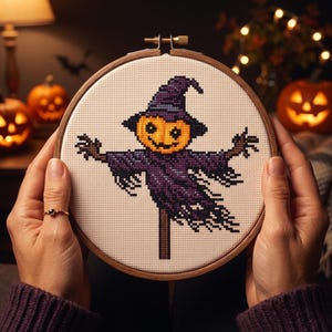 Puede incluir: Un bastidor de bordado circular de madera que contiene un patr&oacute;n de punto de cruz de un espantap&aacute;jaros con cabeza de calabaza, con un sombrero puntiagudo morado y ropa morada harapienta sobre un fondo de tela blanca.