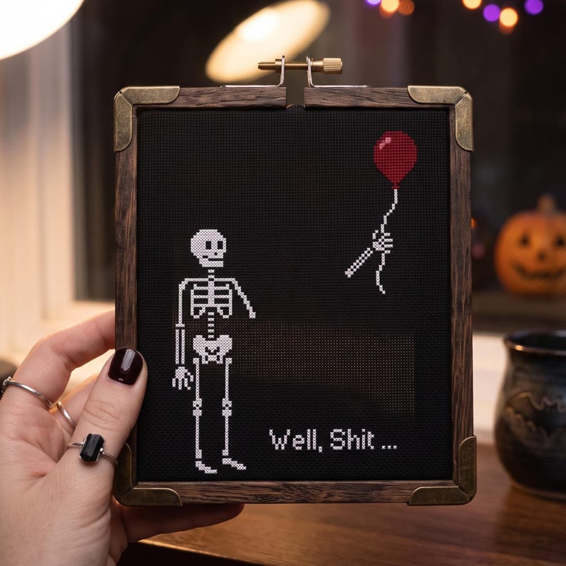 K&ouml;nnte beinhalten: Ein quadratisches Kreuzstich-Stickbild in einem dunklen Holzrahmen mit Messing-Eckbeschl&auml;gen. Der schwarze Stoff zeigt ein wei&szlig;es Skelett, einen roten Luftballon in einer Skeletthand und den wei&szlig;en Sticktext, der lautet, Well, Shit...