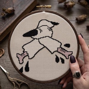 May include: A circular wooden embroidery hoop containing a cross-stitch pattern on cream-coloured fabric. The design features a stylised black and white figure with pink bone-like appendages and scattered black shapes against the fabric background.