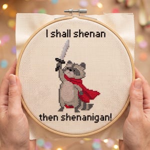 Pu&ograve; includere: Un ricamo a punto croce in un telaio rotondo di legno raffigurante un procione grigio che indossa un mantello rosso e impugna una spada argentata. Il testo sopra recita I shall shenan e quello sotto then shenanigan! su tessuto bianco.