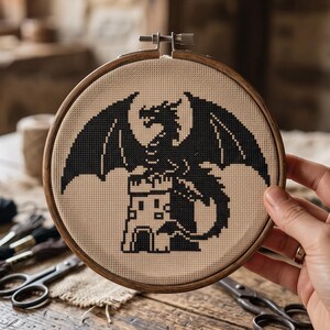 Peut inclure: Un tambour &agrave; broder circulaire en bois contenant un ouvrage au point de croix sur tissu beige. Le motif repr&eacute;sente la silhouette noire d'un dragon aux grandes ailes perch&eacute; sur une tour de ch&acirc;teau, r&eacute;alis&eacute; en style pixelis&eacute;.