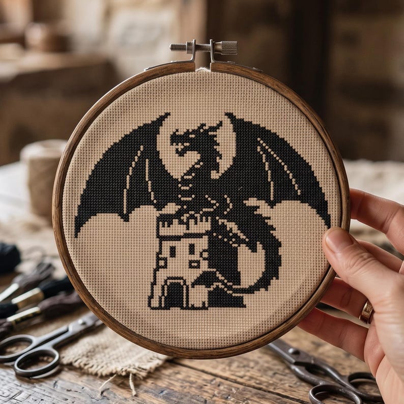 Peut inclure: Un tambour &agrave; broder circulaire en bois contenant un ouvrage au point de croix sur tissu beige. Le motif repr&eacute;sente la silhouette noire d'un dragon aux grandes ailes perch&eacute; sur une tour de ch&acirc;teau, r&eacute;alis&eacute; en style pixelis&eacute;.