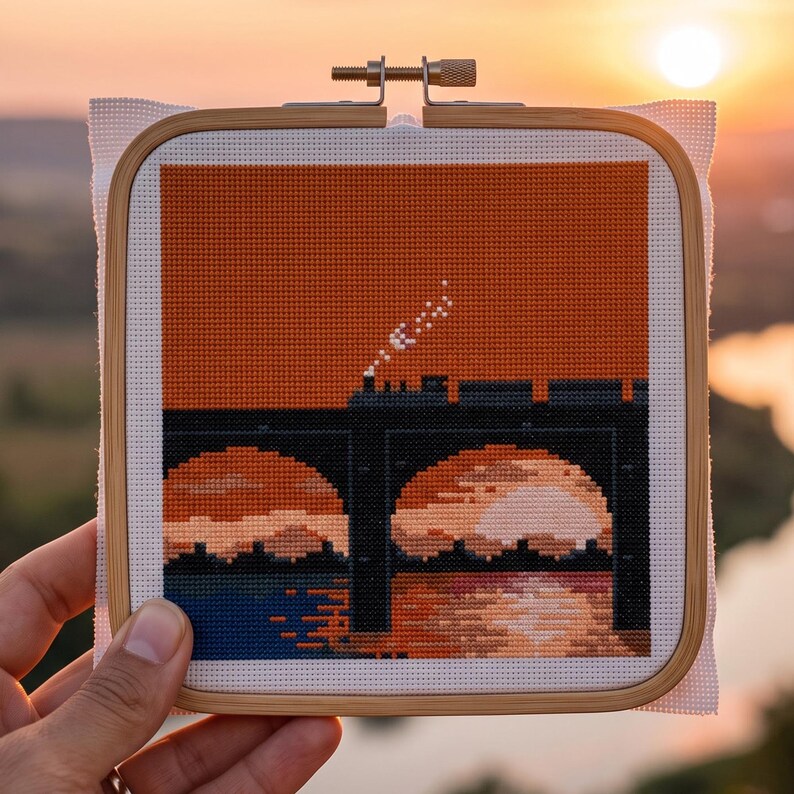 Puede incluir: Un bordado de punto de cruz cuadrado en un bastidor de madera que muestra la silueta de un tren oscuro cruzando un puente sobre el agua al atardecer. El dise&ntilde;o utiliza hilos naranjas, negros y azules sobre tela Aida blanca con vapor.