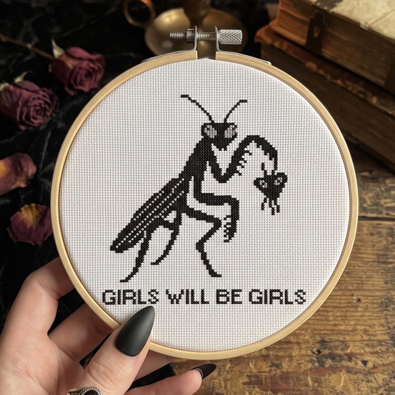 May include: A circular wooden embroidery hoop containing white fabric with a black cross-stitch design of a praying mantis holding a small heart. The text "GIRLS WILL BE GIRLS" is stitched in black capital letters below the insect figure.